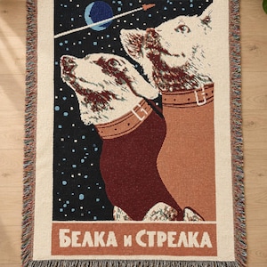 May include: A rectangular woven throw blanket featuring a space-themed graphic of two dogs in collars against a dark blue starry sky with a blue planet and rocket. The text "БЕЛКА и СТРЕЛКА" is printed in white at the bottom on a terracotta background.