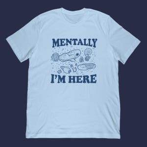 May include: Pale blue t-shirt with a navy blue graphic. The graphic features the text "MENTALLY I'M HERE" above and below illustrations of sea creatures. The illustrations include a whale, jellyfish, and other marine life.