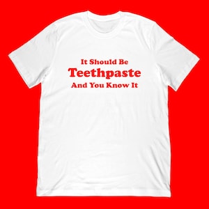 Op de afbeelding: Wit t-shirt met de rode tekst "It Should Be Toothpaste And You Know It". Het shirt heeft een ronde hals en korte mouwen. De achtergrond is effen rood.