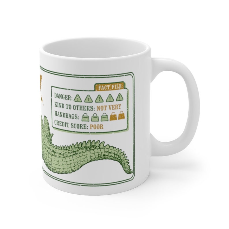 May include: White ceramic mug with a green alligator illustration and a fact file about the animal. The fact file says "Danger: AAAA", "Kind to others: Not very", "Handbags:  ", "Credit score: Poor".