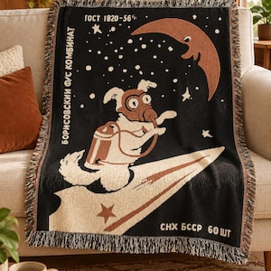 May include: A black woven throw blanket featuring a white dog wearing a space helmet and oxygen tank, riding a paper airplane through a starry sky with a crescent moon. Text includes "ГОСТ 1820-56", "БОРИСОВСКИЙ Ф/С КОМБИНАТ", and "СНХ БССР 60 ШТ".