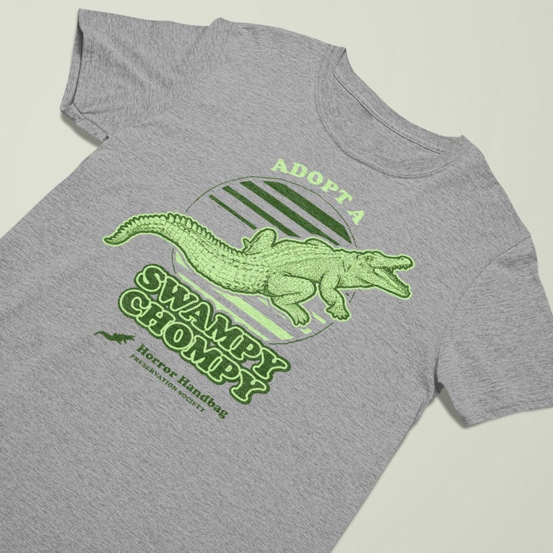 May include: A grey t-shirt with a green alligator graphic and the text "Adopt a Swampy Chompy" and "Horror Handbag Preservation Society".