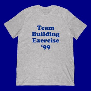 Puede incluir: Camiseta gris jaspeado con el texto azul "Team Building Exercise '99". La camiseta de manga corta, con cuello redondo, está hecha de un material suave. El texto está centrado en la parte delantera.