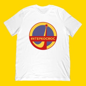 May include: White t-shirt featuring a circular graphic with a blue background, a yellow arc, and a red star. The graphic includes the Russian text "ИНТЕРКОСМОС" in red. The shirt has short sleeves and a crew neck.