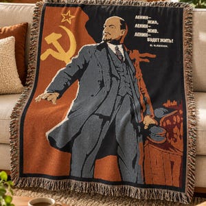 May include: A woven throw blanket featuring a graphic of Vladimir Lenin in a dark suit, a yellow hammer and sickle symbol, and the Cyrillic text "ЛЕНИН- ЖИЛ, ЛЕНИН- ЖИВ, ЛЕНИН- БУДЕТ ЖИТЬ! В. И. ЛЕНИН." The blanket has a fringed edge and a color palette of black, orange, and tan.