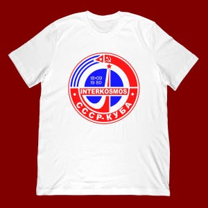 May include: White t-shirt featuring a circular graphic with the text "INTERKOSMOS" and Cyrillic text. The graphic includes red and blue elements, a star, and the dates "18-09 19 80".
