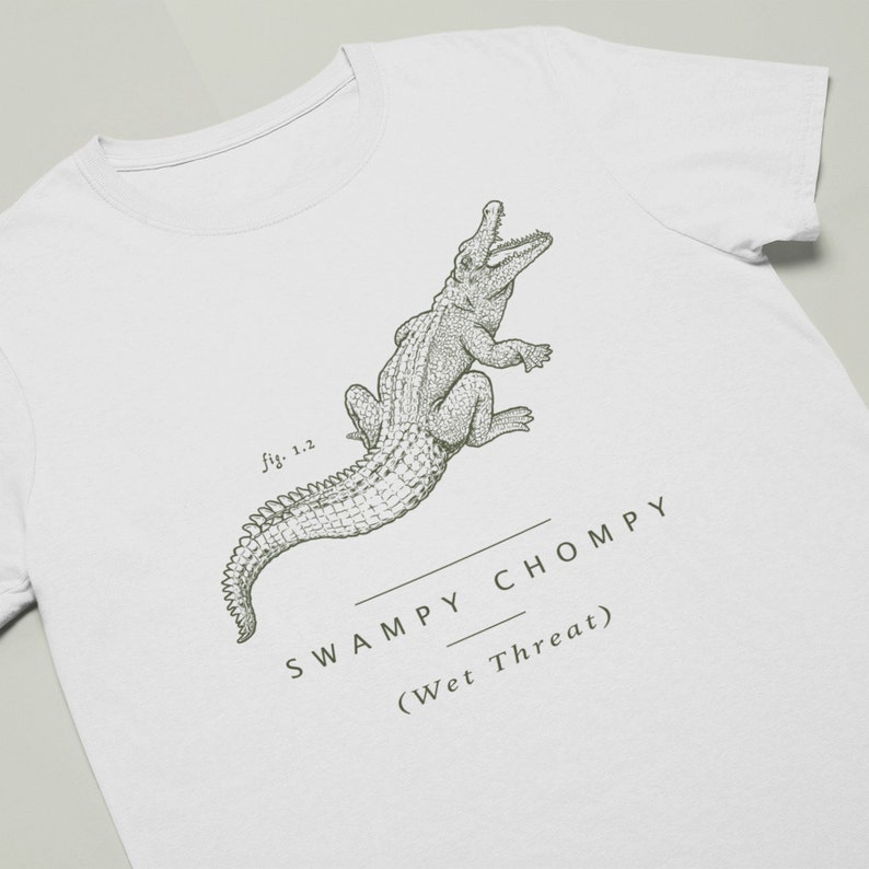 May include: White t-shirt featuring a detailed illustration of an alligator with the text "SWAMPY CHOMPY (Wet Threat)". The design is in a muted green color, with the text and illustration centered on the shirt.