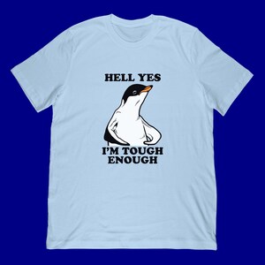 Può includere: T-shirt azzurra con un'illustrazione di pinguino e la scritta "HELL YES I'M TOUGH ENOUGH". Il pinguino è bianco e nero con un becco arancione. La maglietta ha un girocollo e maniche corte.