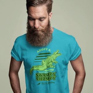 May include: A turquoise blue t-shirt featuring a green alligator graphic and the text "Adopt a Swampy Chompy" in a retro font. The text "Horror Handbag Presents" is also included in smaller font.