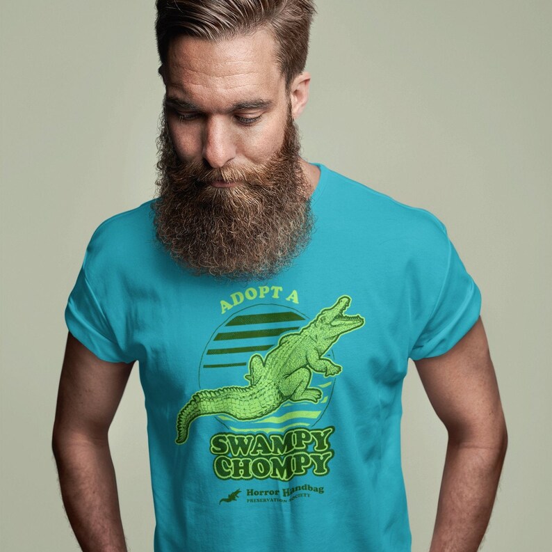May include: A turquoise blue t-shirt featuring a green alligator graphic and the text "Adopt a Swampy Chompy" in a retro font. The text "Horror Handbag Presents" is also included in smaller font.