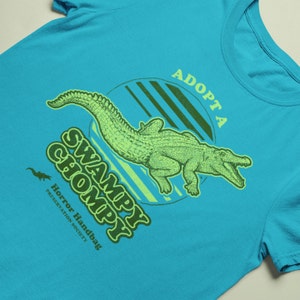 May include: A turquoise blue t-shirt featuring a green alligator graphic and the text "Adopt a Swampy Chompy" in green and yellow lettering. The text "Horror Handbag Preservation Society" is also printed in smaller lettering below the alligator.