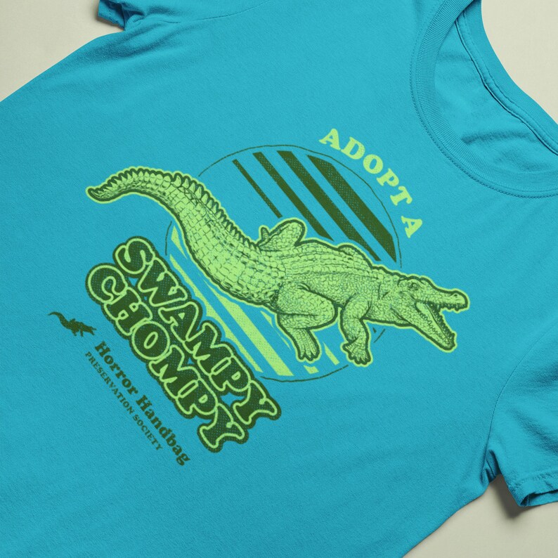 May include: A turquoise blue t-shirt featuring a green alligator graphic and the text "Adopt a Swampy Chompy" in green and yellow lettering. The text "Horror Handbag Preservation Society" is also printed in smaller lettering below the alligator.