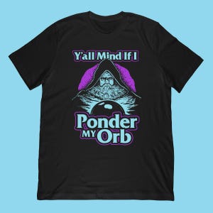 Puede incluir: Camiseta negra con un diseño gráfico de un mago encapuchado, una bola de cristal y el texto "Y'all Mind If I Ponder My Orb" en morado y azul. El diseño presenta una figura barbuda con una expresión seria.