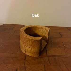 May include: A handcrafted, semi-circular wooden coaster made of oak. The coaster has a natural wood grain pattern and a smooth, curved design. The word "Oak" is printed on the top of the coaster. This coaster is designed to hold a glass or mug.