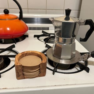 May include: A stainless steel stovetop espresso maker and a wooden holder with a stack of coffee filters sit on a white stove. An orange kettle with a black handle is also on the stove.