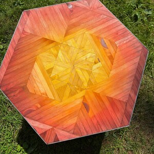 May include: Hexagonal wooden table with a vibrant, geometric pattern. The tabletop features a gradient of colors, transitioning from red to orange to yellow. The table has a silver metal edge and is set on a grassy surface.