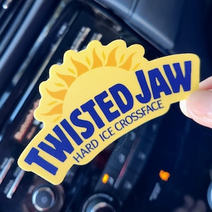 May include: A yellow sticker with the text "TWISTED JAW" in blue, with a yellow sun graphic. The sticker also includes the text "HARD ICE CROSSFACE". The sticker is held up against a dark background.