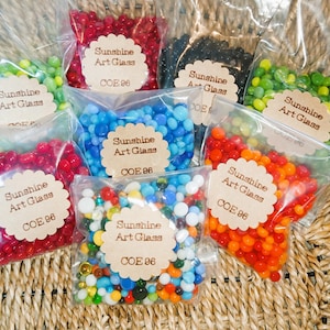 Puede incluir: Variedad de cuentas de vidrio de colores en bolsas de plástico transparente. Cada bolsa tiene una etiqueta con la inscripción "Sunshine Art Glass COE 96". Las cuentas son de varios colores, incluyendo rojo, azul, verde y naranja. Las bolsas están dispuestas sobre una superficie tejida.