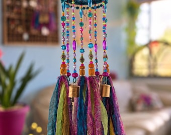 May include: A hanging mobile featuring a circular frame adorned with vibrant purple, blue, and green beads. Strands of colorful beads extend downward, interspersed with metallic bells and long, flowing fabric ribbons in shades of lime, teal, and magenta.