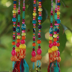 Op de afbeelding: Een kleurrijke kralenwindgong met een turquoise top en gouden bellen. De kralen zijn geregen op meerdere strengen en omvatten tinten roze, oranje, geel, blauw en groen.