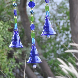 Puede incluir: Tres campanas de vidrio azul colgando de una cuerda de cuentas azules, verdes y blancas. Las campanas están suspendidas de una varilla de madera.
