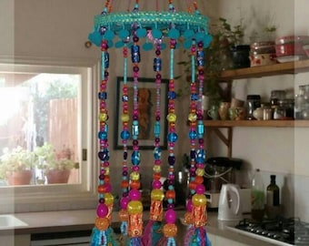 May include: A colorful beaded wind chime with a turquoise top and hanging strands of beads in various colors. The strands end with gold-colored bells and colorful fabric ribbons. The wind chime is hanging in a kitchen.