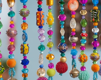 Op de afbeelding: Een levendig kralengordijn met strengen kleurrijke kralen in verschillende vormen en maten. De kralen zijn in tinten blauw, roze, oranje, geel, groen en goud. Sommige kralen hebben ingewikkelde patronen, andere een getextureerd uiterlijk.