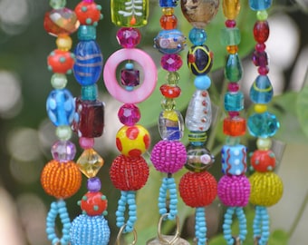 May include: A colorful beaded wind chime with bells. The beads are made of glass and are in various colors, including red, blue, green, yellow, and pink. The bells are made of brass and are attached to the bottom of the chime.