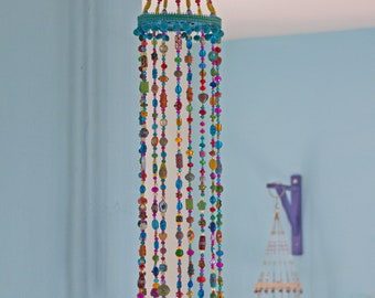 May include: A colorful beaded wind chime with bells hanging from the bottom. The chime is made of various colors of beads, including blue, green, red, yellow, and orange. The chime is hanging from a blue ceiling mount.