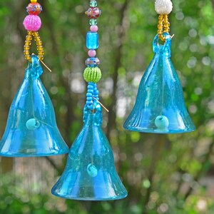 Puede incluir: Tres campanas de vidrio azul colgando de cuerdas con cuentas de colores. Las campanas son transparentes y tienen un ligero brillo iridiscente.