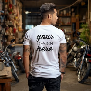May include: A white t-shirt with the text "your DESIGN here" printed on the back. The shirt is being worn by a person with tattoos on their arms. The person is standing in a garage with motorcycles in the background.
