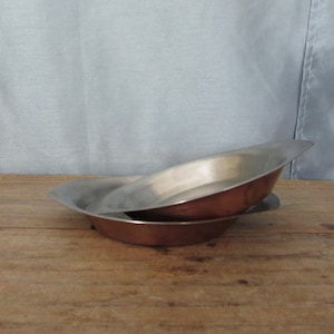 May include: Two metal bowls, one stacked on top of the other. The top bowl is silver, and the bottom bowl is copper-colored. The bowls are on a wooden surface, with a light blue background.