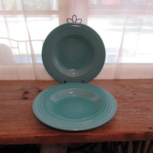 May include: Two turquoise-colored ceramic bowls with concentric ring detailing. One bowl is displayed on a black metal stand. The bowls are on a rustic wooden surface, with a window and sheer curtains in the background.