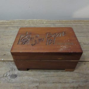 May include: A rectangular wooden music box with a carved treble clef and musical notes design. The words "Beat It!" are carved into the top. The box is a warm brown color and sits on small feet.