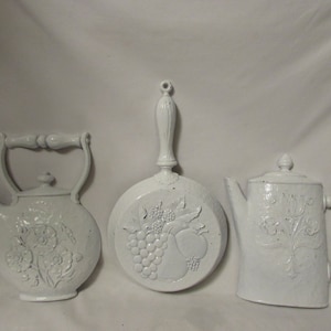May include: Three white decorative kitchen wall hangings. The set includes a teapot, a frying pan with a fruit design, and a teapot with a lid. Each piece has a textured surface and is painted white.