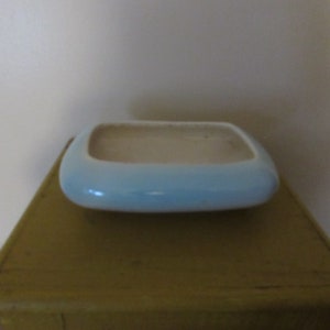 May include: A small, rectangular, light blue ceramic planter with rounded corners. The interior is a cream color. The planter is sitting on a yellow-brown surface. This decorative piece is suitable for small plants or succulents.
