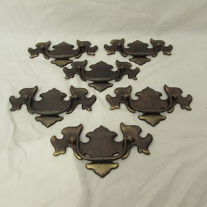 May include: Set of six antique bronze drawer pulls. Each pull features a decorative design with a curved handle and ornate detailing. The pulls are a warm bronze color with hints of gold, perfect for vintage furniture restoration.