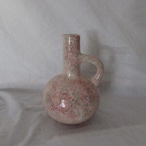 May include: A speckled ceramic pitcher with a round base, a handle, and a narrow spout. The pitcher is primarily pink with green and red speckles. The handle is curved and attached to the body of the pitcher.