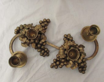 May include: A pair of vintage brass candle holders. Each holder features a curved arm with a candle cup and is adorned with grape clusters and leaves. The candle holders are a warm gold color and are set against a white background.