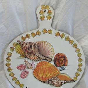May include: A white, round cutting board with a handle, decorated with illustrations of various seashells in shades of brown, orange, and pink. The words "BERMUDA SHELLS" are printed in black at the bottom.