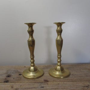 May include: Two brass candlesticks with a classic design. Each candlestick has a round base, a slender stem with decorative detailing, and a flared top to hold a candle. The candlesticks are made of shiny, polished brass.
