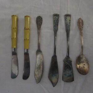 May include: A collection of antique silverware, including two butter knives with yellow bamboo-style handles, two butter spreaders, and a spoon. The silverware is arranged on a white surface.