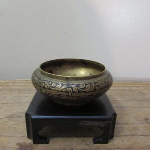 May include: A brass bowl with a dark blue and gold floral design sits on a black wooden stand. The bowl has a rounded shape and a wide opening. The floral pattern is intricate and detailed, adding a touch of elegance.
