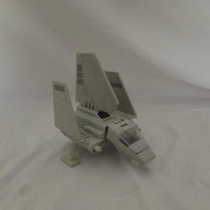 May include: A light gray toy spaceship with angled wings and a cockpit. The wings are positioned at different angles, and the body has detailed panel lines. The toy is made of plastic and sits on a small stand.