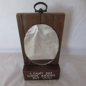 May include: A wooden framed mirror with a dark brown finish. The mirror has an oval shape and a metal handle at the top. The base of the frame has the text "I CAN'T SEE LOVIN' ANYONE BUT YOU" in white.