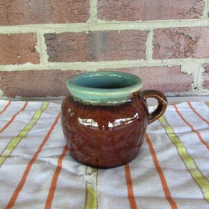 May include: A brown and green ceramic mug with a handle. The mug has a glazed finish and is sitting on a striped towel.