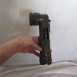 May include: Olive green, right-angle flashlight. The flashlight has a cylindrical body with a textured grip and a pivoting head. It features a clip and a switch on the side. The lens is at the end of the head.