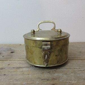 May include: A round, antique brass box with a handle and clasp. The box is cylindrical with a lid and a handle on top. The clasp is located on the front of the box. The box is sitting on a wooden surface.