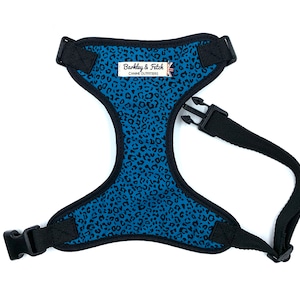 May include: A blue and black dog harness with a leopard print pattern. The harness has a black adjustable strap and a buckle closure. The harness has a label that reads "Barkley & Fetch Canine Outfitters".