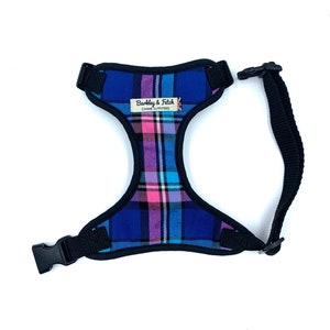 May include: A blue, pink and white tartan dog harness with black adjustable straps and a buckle fastening. The harness has a label that reads "Barkley & Fitch Canine Outfitters".